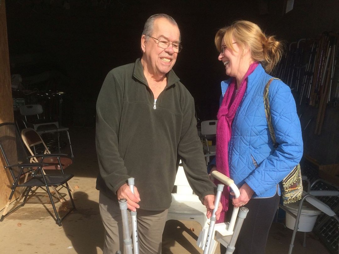 A Lions volunteer and a smiling community member with a loaned walker and commode chair outside the club's medical equipment shed